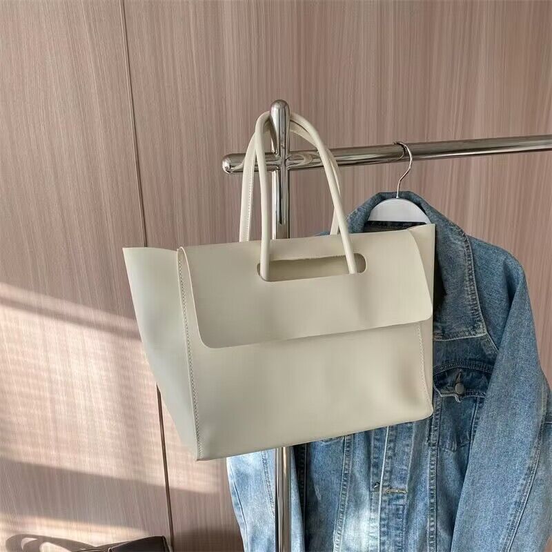 Shoulder bag with large capacity in stylish design, light beige color, hanging on a garment rack with a denim jacket.